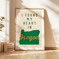 A framed poster with the text "I FOUND MY HEART IN OREGON" is displayed on a wooden floor next to a stack of books.