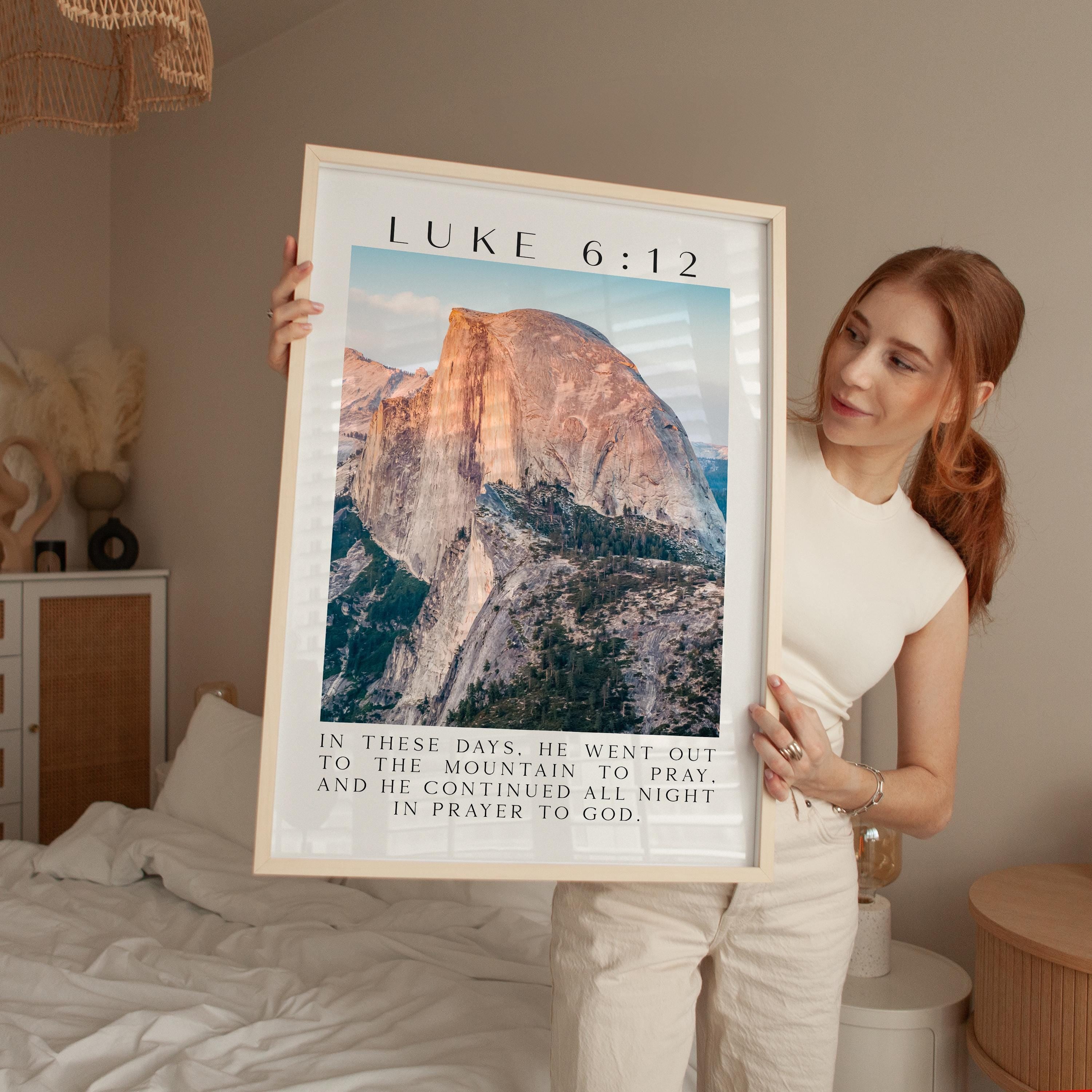 A woman is holding up a framed poster with a mountain landscape and text, which appears to be a biblical verse from the book of Luke.