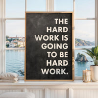 May include: A rectangular framed wall art piece featuring a dark charcoal gray background with a textured, chalkboard-like appearance. The white, sans-serif text reads: THE HARD WORK IS GOING TO BE HARD WORK. The frame is a light-toned natural wood.