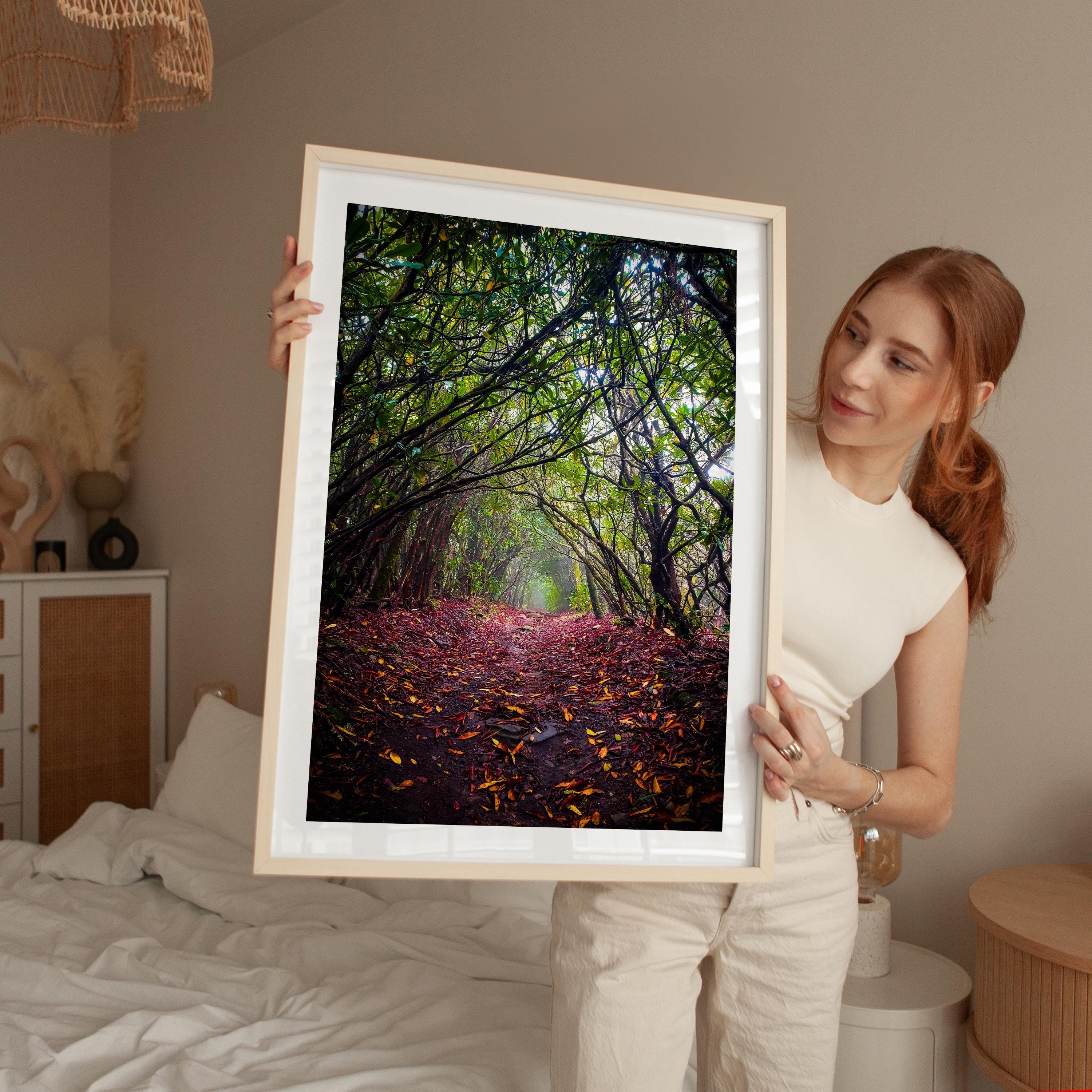A woman holds a framed photograph of a forest path with fallen leaves, standing in a bedroom with a bed and other furniture visible in the background.