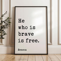 A framed poster with the text "He who is brave is free" by Seneca, standing on a wooden floor against a white wall.