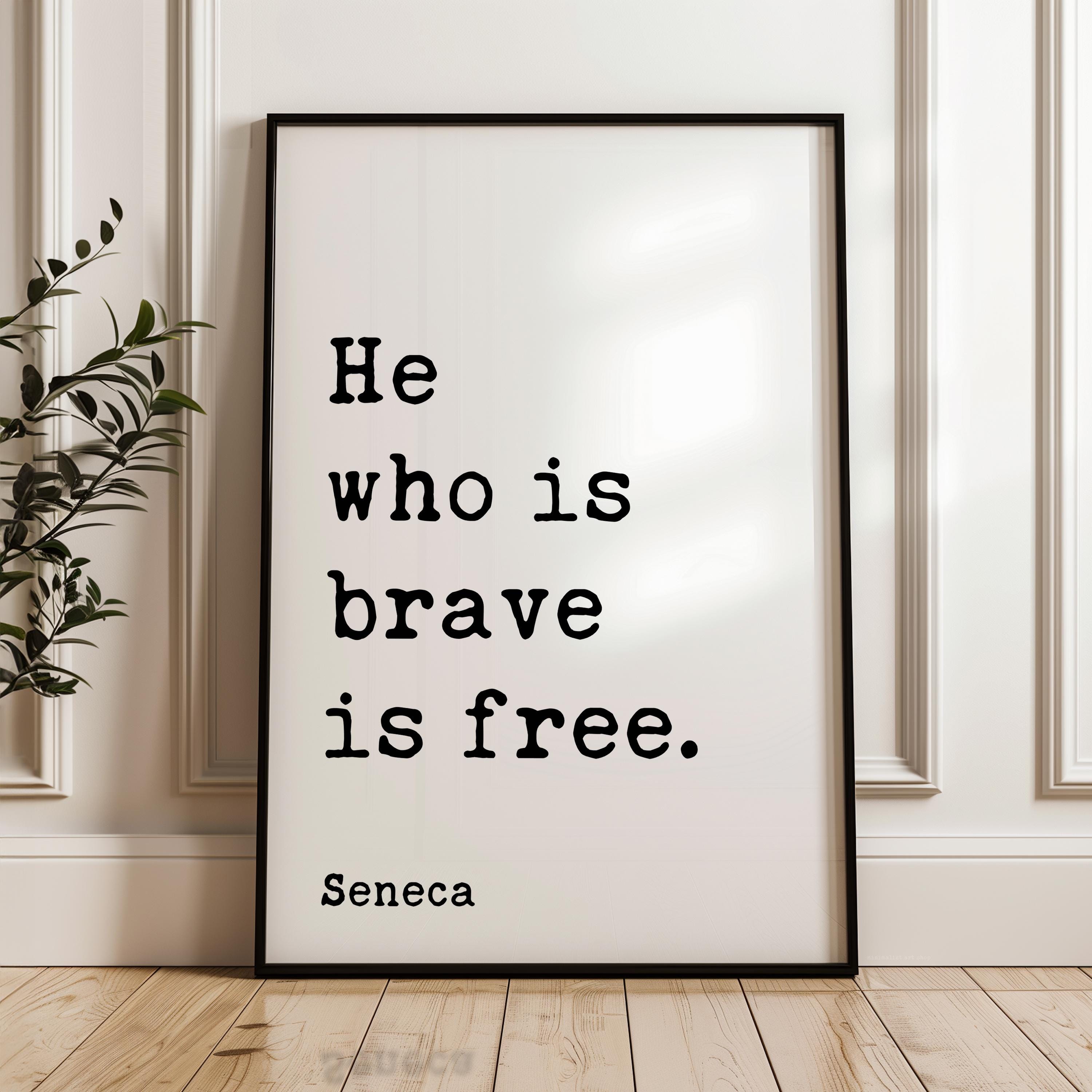 A framed poster with the text "He who is brave is free" by Seneca, standing on a wooden floor against a white wall.