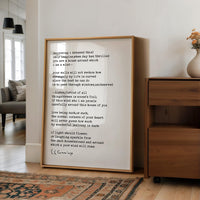 a framed poem with a wooden frame, placed on a wooden floor in a room with a vase of flowers and a couch.