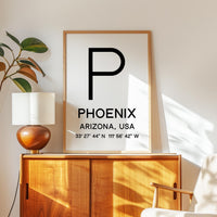 a framed poster with the text "PHOENIX, ARIZONA, USA" and a coordinate system, along with a lamp and a plant on a wooden cabinet.