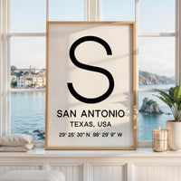 May include: A framed vertical wall art print featuring a large black letter S at the top, followed by the text SAN ANTONIO, TEXAS, USA, and the geographic coordinates 29° 25' 30" N 98° 29' 9" W printed in black on a white background.