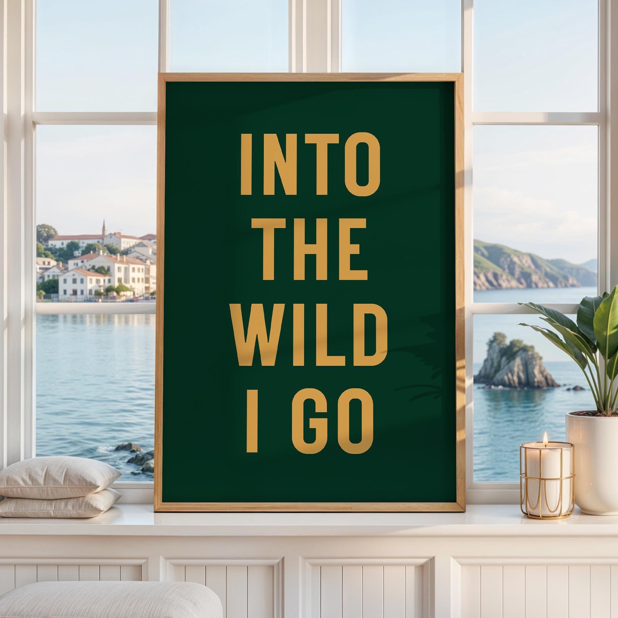 May include: A rectangular wall art print in a light wood frame featuring a dark green background with the gold text INTO THE WILD I GO centered in a bold sans-serif font. The print is displayed against a window with a view of a coastal town.