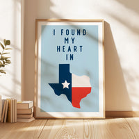 A framed poster with the text "I FOUND MY HEART IN" and a graphic of the state of Texas, placed on a wooden floor next to a stack of books.